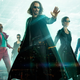 Keanu Reeves, wearing sunglasses and a long black coat, poses with his hand out in front of the the Matrix Resurrections cast.